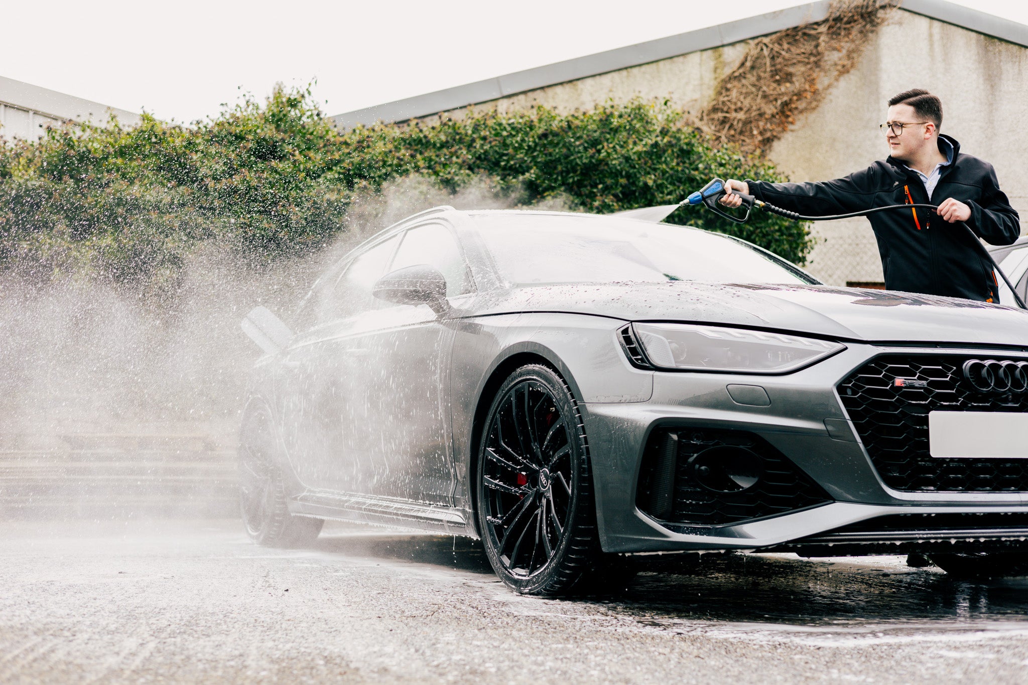 A man wearing glasses and a black jacket is performing a car wash on a sleek grey Audi S-series using a high-pressure hose. Water sprays across the vehicle, creating a dynamic effect as foam and droplets scatter. This car cleaning process is part of a thorough car detailing routine, ensuring the car’s exterior remains spotless. The black alloy wheels and aggressive front grille enhance its sporty appearance. The scene takes place outdoors with greenery and a modern building in the background.