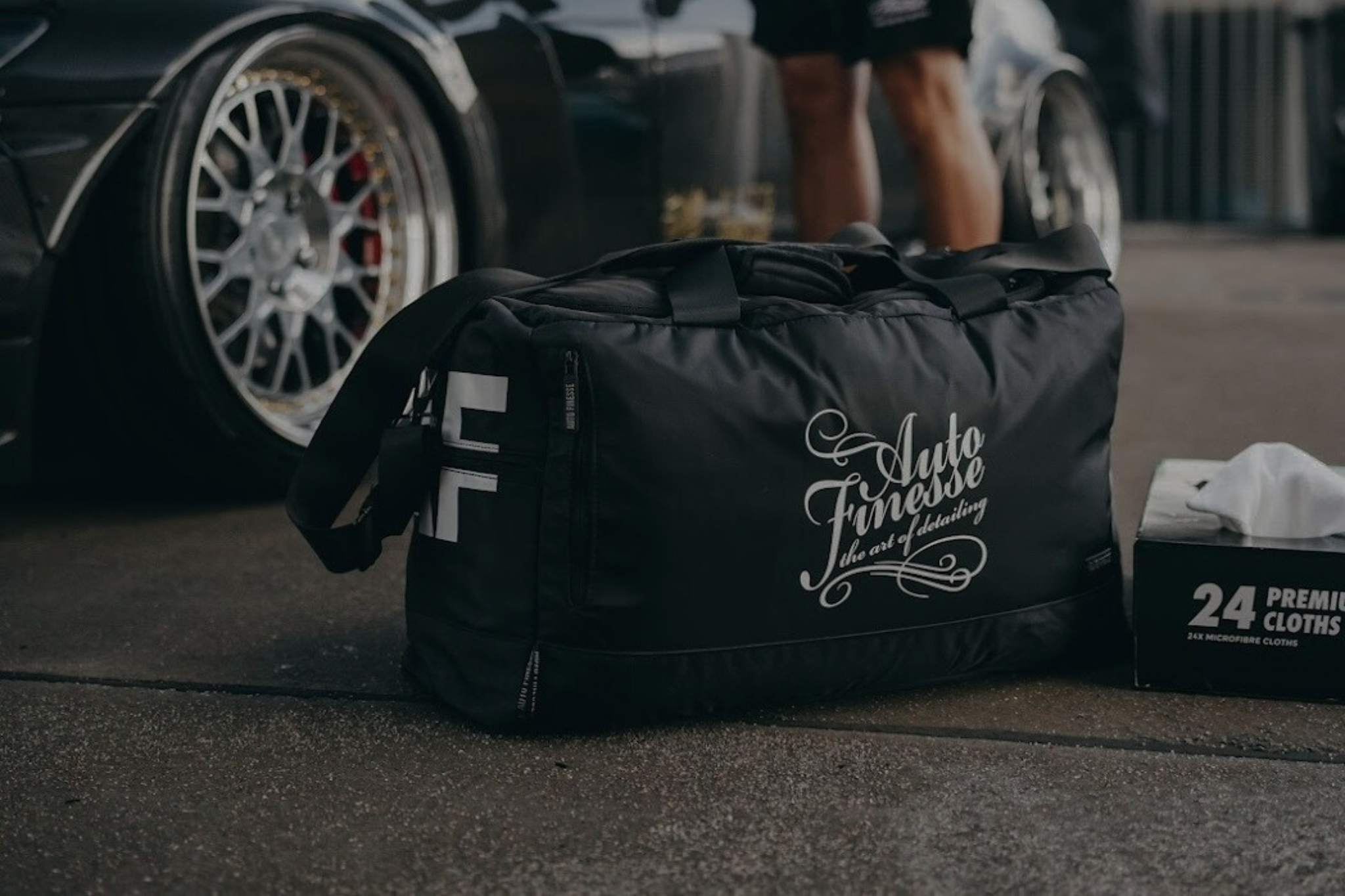 Black duffel bag with 'Auto Fitness' branding on a concrete surface with a car in the background.
