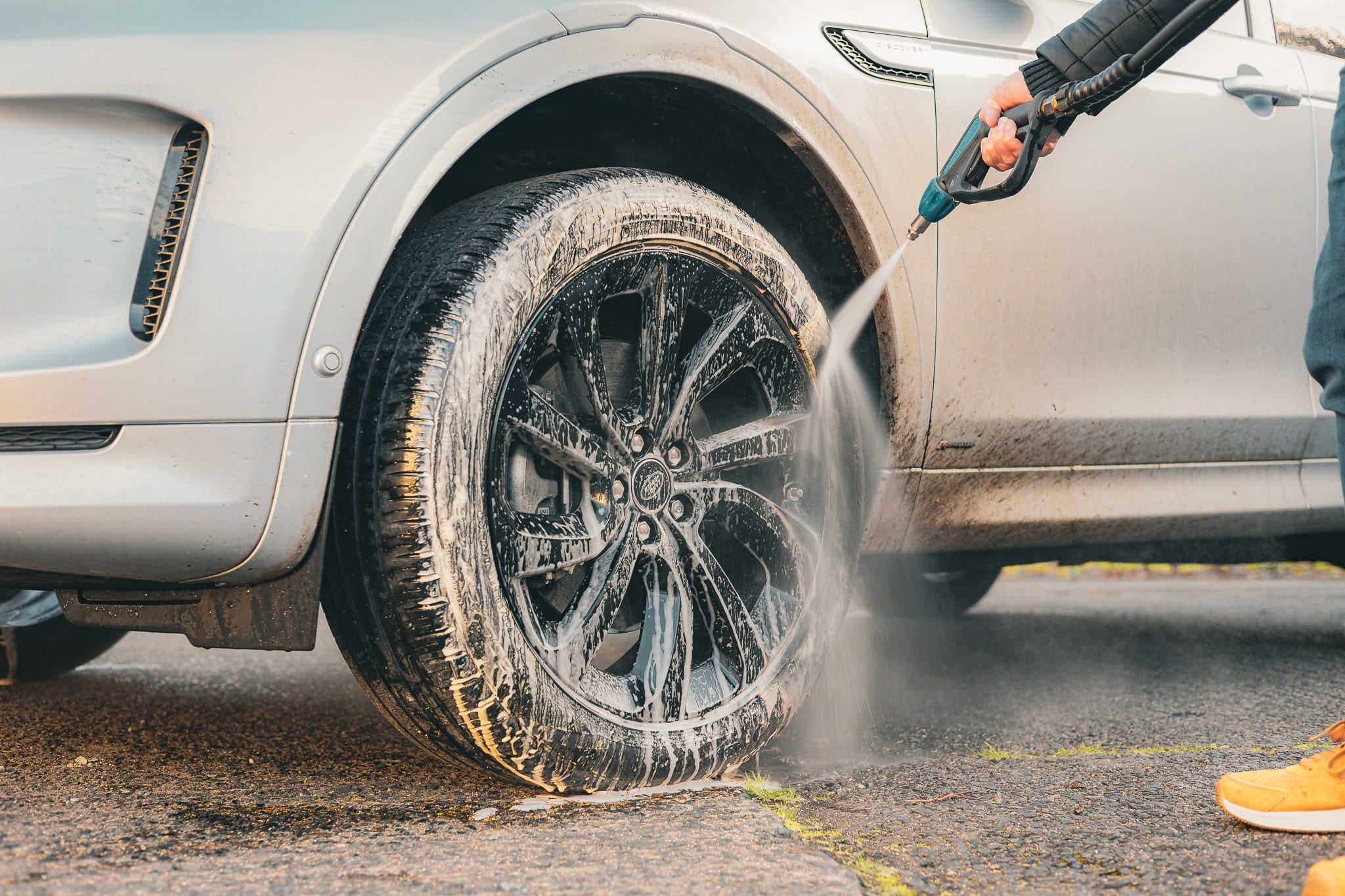 Pressure washer rinsing a foam-covered alloy wheel during professional car detailing. Safe wheel cleaning removes brake dust, tyre dressing residue, road grime and traffic film before applying wheel protection for a spotless finish.