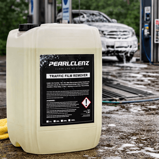 A large white plastic jerrycan of Pearlclenz Traffic Film Remover with a black label, placed on a wet concrete surface at a car wash with a soapy car blurred in the background.